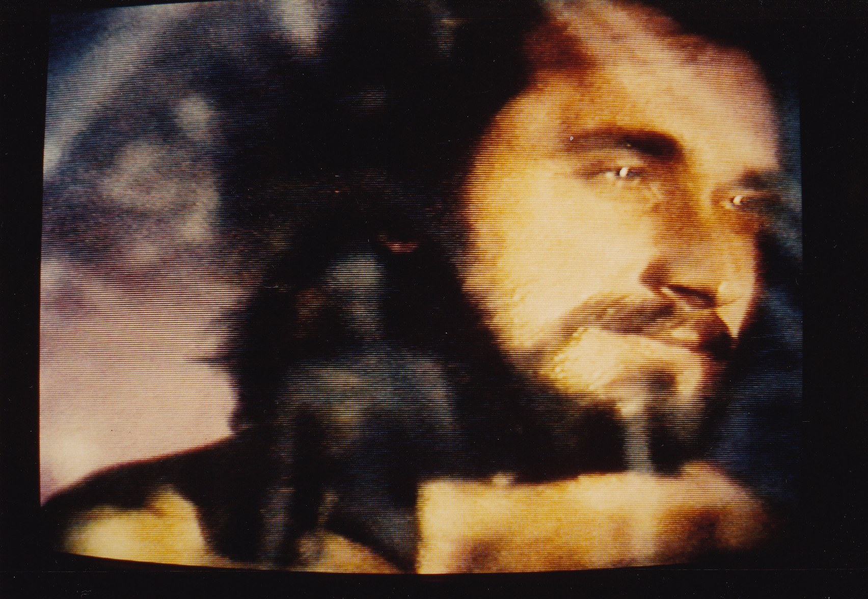 Mann mit dunklem Bart und Schnurrbart, der eine weiße Hemd trägt und direkt in die Kamera schaut mit einem ernsten Gesichtsausdruck vor einem dunklen Hintergrund.