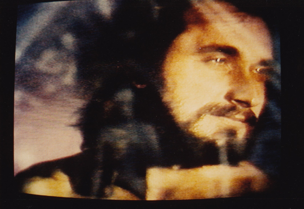 Mann mit dunklem Bart und Schnurrbart, der eine weiße Hemd trägt und direkt in die Kamera schaut mit einem ernsten Gesichtsausdruck vor einem dunklen Hintergrund.