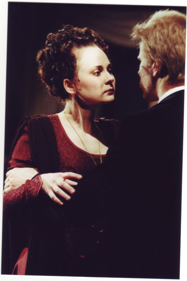 Eine Frau in einem roten Kleid mit einer Kette steht neben einem Mann in einem schwarzen Anzug, der sie liebevoll ansieht, vor einem dunklen Hintergrund.