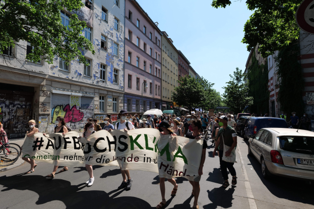 Eine Gruppe von Menschen marschiert auf einer Straße in Berlin, hält ein Transparent hoch, mit parkenden Autos auf der rechten Seite, einem Fahrradfahrer auf der linken Seite und Gebäuden, Bäumen und einer Schautafel im Hintergrund unter einem sichtbaren Himmel.