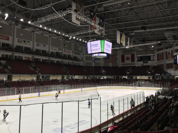 Ein Eishockeyspiel in einer großen Arena mit einem zentralen Netz, Zuschauern auf beiden Seiten und Bannern an den Wänden unter einer Decke mit Lampen und Trägern, identifiziert als die St. Louis Blues Arena.