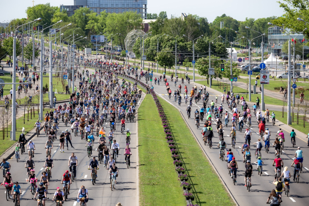 Eine große Gruppe von Menschen auf Fahrrädern fährt eine Straße entlang, die von Grünflächen, Verkehrsschildern, Schildern, Fahrzeugen, Bäumen, Gebäuden und einem klaren blauen Himmel gesäumt ist.