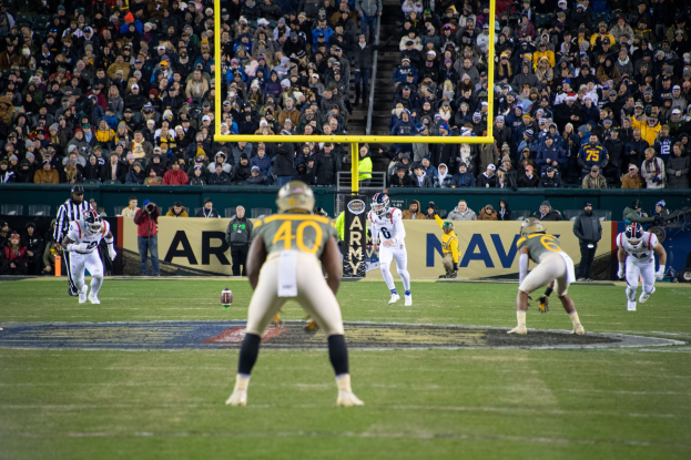 Fußballspieler in Sportkleidung und Helmen konkurrieren auf einem Feld, mit Zuschauern auf den Tribünen und Stadionwerbung im Hintergrund.