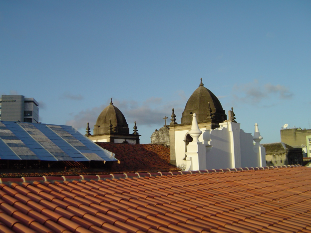 Stadtpanorama mit Gebäuden im Vordergrund, Solarpaneele auf einem Dach und einem blauen Himmel im Hintergrund.
