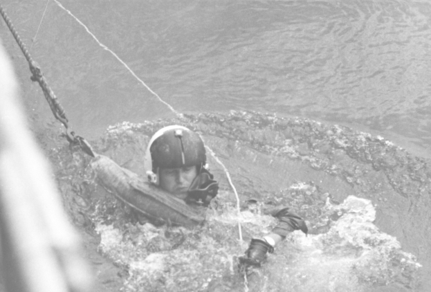 Schwarzes Bild eines Retters in einem Neoprenanzug und Helm, der in flachem Wasser treibt und an einem Seil befestigt ist.