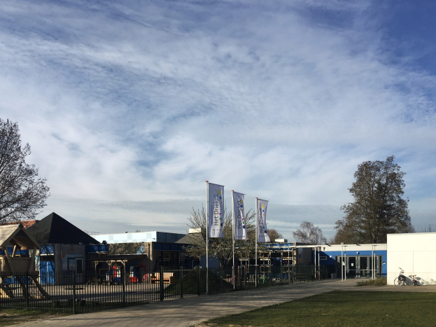 Großes Gebäude mit einem grasbewachsenen Bereich davor, umgeben von einem Metallzaun, mit einem Weg, Bäumen, Pfosten, Bannern, einem geparkten Fahrrad, nahen Häusern und einem bewölkten Himmel, als neuer Spielplatz an der Schule identifiziert.
