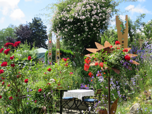 Ein Garten auf der Chelsea Flower Show mit bunten Blumen und Pflanzen, einem Tisch mit einer Decke umgeben von Stühlen, Bäumen und einem bewölkten Himmel im Hintergrund.