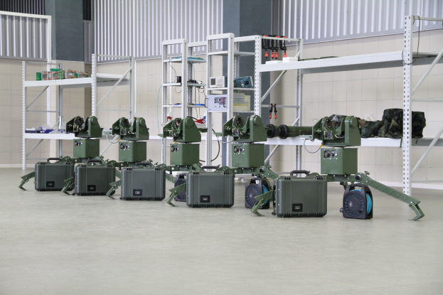 Eine Gruppe von Milit├Ąrrobotern auf einem Boden mit Koffern und Gegenst├Ąnden auf Gestellen, in der N├Ąhe einer Wand und eines Gel├Ąnders als Teil eines Verteidigungssystems.