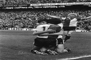 Drei Fussballspieler feiern ein Tor auf dem Boden, umgeben von einer jubelnden Menge in einem schwarz-weissen Stadion.
