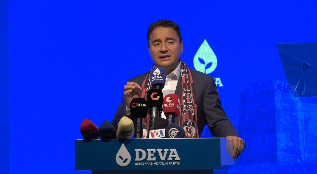 Ein Mann in Mantel und Schal steht an einem Pult mit Mikrofonen und spricht zu einem Publikum; auf dem Pult ist eine Tafel mit dem Text 'deva-Gesch├Ąftsführer sagt, es sei keine gute Idee für die Zukunft', mit einem Bildschirm und einem Gegenstand im Hintergrund.