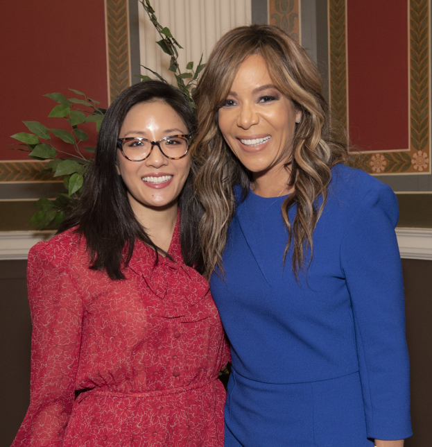 Zwei Frauen in bunten Kleidern lächeln gemeinsam auf einem Event, wobei eine eine Brille trägt und im Hintergrund eine Pflanze und ein Vorhang zu sehen sind.