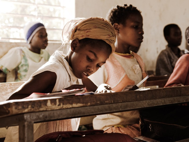 Eine Gruppe von Kindern sitzt an Tischen in einem Klassenzimmer, mit Büchern und Taschen auf den Bänken vor ihnen und einem Fenster und einer Wand im Hintergrund.