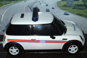 Ein weißer Polizeiwagen steht im Vordergrund mit mehreren fahrenden Fahrzeugen auf der Straße dahinter, flankiert von Gras und Bäumen auf beiden Seiten in einer ländlichen Umgebung.