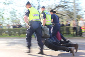 Gruppe von Polizisten in Mützen, die einen Mann mit einer Tasche die Straße entlangschieben.