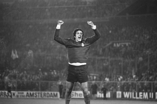 Schwarzes und weißes Foto eines Mannes, der auf einem Fußballfeld mit erhobenen Armen feiert, umgeben von einer Menge, einem Zaun und Bannern.