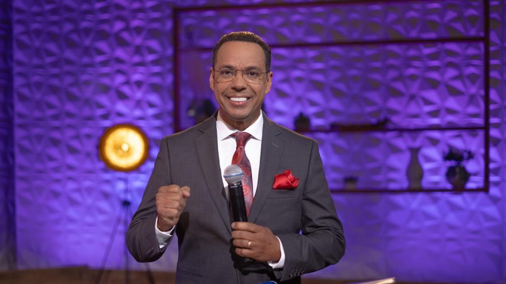 Stephen Colbert, Moderator von "The Daily Show with Trevor Noah", steht auf einer Bühne und hält ein Mikrofon in der Hand, trägt einen Anzug, eine Krawatte und eine Brille, während im Hintergrund ein Scheinwerfer und ein Ständer zu sehen sind.