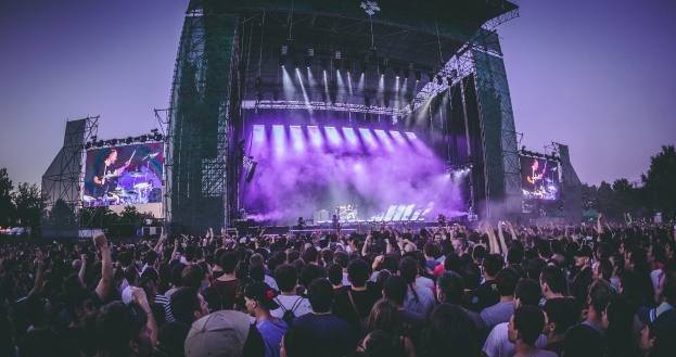 Eine große Menge bei einem Musikfestival schaut auf einer hell beleuchteten Bühne mit Instrumenten und Bildschirmen, die vor einem Gebäude, Bäumen und einem klaren blauen Himmel steht.
