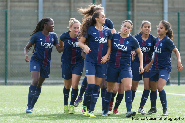 Gruppe junger Frauen beim Fußballspielen auf einem Rasenplatz mit Maschendrahtzaun und einer Wand im Hintergrund, Text in der rechten unteren Ecke lautet 'Paris Saint-Germain Frauenfußball'.