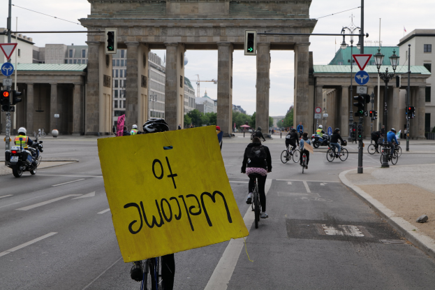 Eine Gruppe von Radfahrern fährt an der Brandenburgertor in Berlin vorbei, einer hält ein gelbes Schild, mit Laternenpfählen, Ampeln, Gebäuden, Bäumen und einem klaren blauen Himmel im Hintergrund.