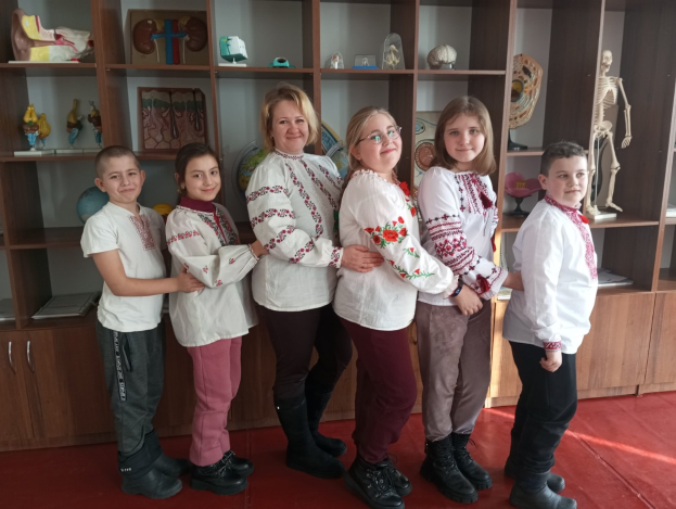 Eine Gruppe von Kindern in traditioneller ukrainischer Kleidung vor einem Bücherregal mit Spielzeug, einem Skelett und anderen Gegenständen, was auf eine ukrainische Schulumgebung hinweist.