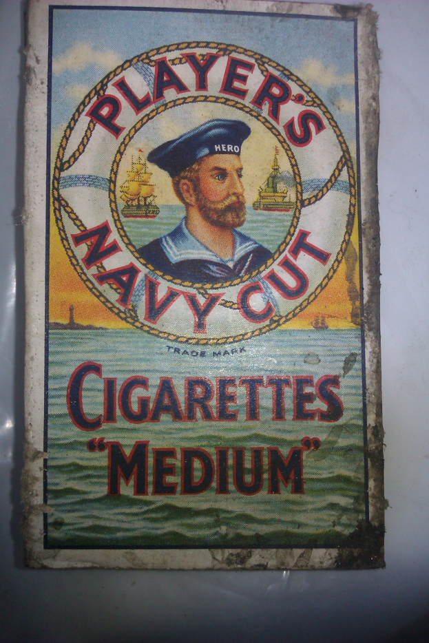 Ein Plakat an einer Wand mit einem Bild einer Person in einer marineblauen Uniform mit weißem Hemd und roter Krawatte, die Arme verschränkt und mit einem strengen Gesichtsausdruck, neben dem Text "Player's Navy Cut Cigarettes Medium" in fetter, schwarzer Schrift mit einem weißen Rand.