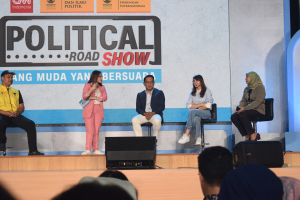 Eine Gruppe von Menschen auf einer Bühne mit einer Frau, die ein Mikrofon hält, in der Mitte, einem sichtbaren Publikum unten und einem Banner mit der Aufschrift 'Politischer Roadshow 2019' im Hintergrund.