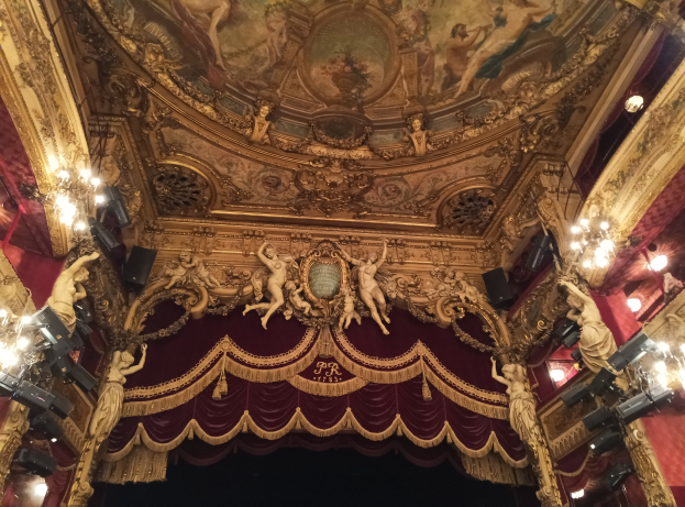 Das Innere eines Theaters mit einem roten Vorhang, Kronleuchtern, Skulpturen, Lichtern und einer Deckenmalerei sowie einer Bühne und einigen Personen unten.