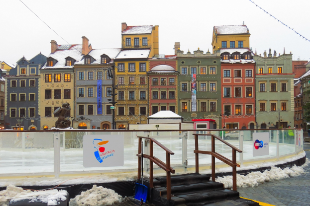 Eislaufbahn in einem Stadtplatz umgeben von Geb├Ąuden, Treppen, Gel├Ąndern, Informationsschildern, schneebedecktem Boden, Laternenmasten, B├Ąumen und einem Himmel als Hintergrund.