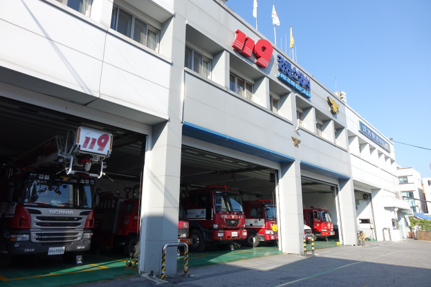 Feuerwache mit mehreren Feuerwehrautos vor der Tür, mit Fenstern, Säulen und Namensschildern, mit Fahnenmasten im Vordergrund und anderen Gebäuden gegen einen klaren blauen Himmel.