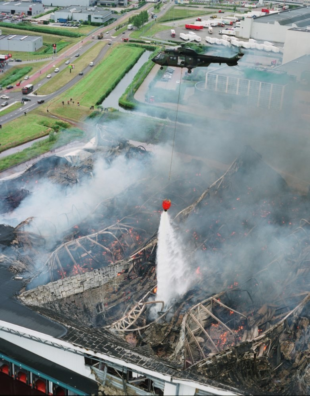 Ein Hubschrauber überfliegt ein großes, durch ein Feuer beschädigten Gebäude mit aufsteigendem Rauch, mit anderen Gebäuden, Fahrzeugen und Bäumen im Hintergrund und Text am unteren Bildrand.