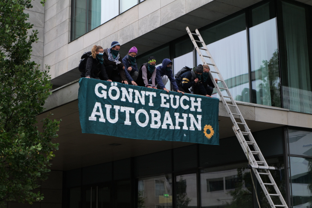 Gruppe von Menschen auf einem Gebäude mit einem Banner, mit einem Baum links und Glasfenstern im Hintergrund, wahrscheinlich protestieren sie für die deutsche Regierung.