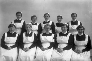 Schwarzes und weißes Foto einer Gruppe von Frauen in weißen Uniformen, die auf einer Bank vor einer Wand sitzen.