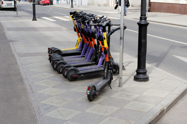 Eine Reihe von Elektro-Scootern, die auf einer Straße neben fahrenden Fahrzeugen, Fußgängern auf dem Gehweg und Gebäuden im Hintergrund geparkt sind.