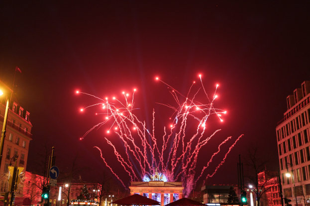 Eine belebte Stadtstraße an einem Neujahrsabend in Berlin, mit Gebäuden, Bäumen, Laternenmasten, Verkehrszeichen, Zeltplanen, Menschen und einem prächtigen Feuerwerk am Himmel.