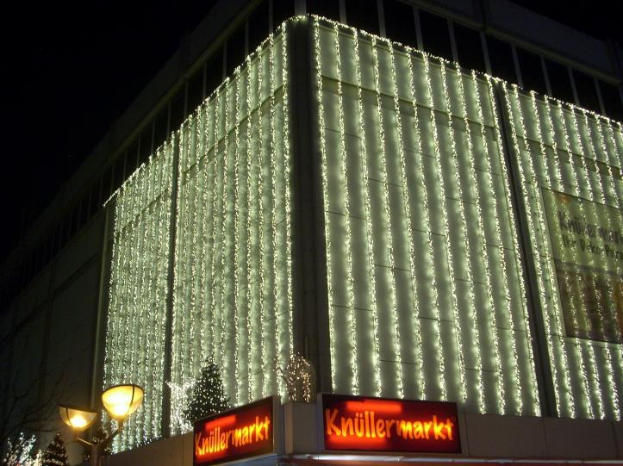 Ein Gebäude in Berlin, Deutschland, geschmückt mit Weihnachtslichtern, umgeben von Bäumen, Laternenmasten und Tafeln mit Text, mit dem Himmel im Hintergrund.
