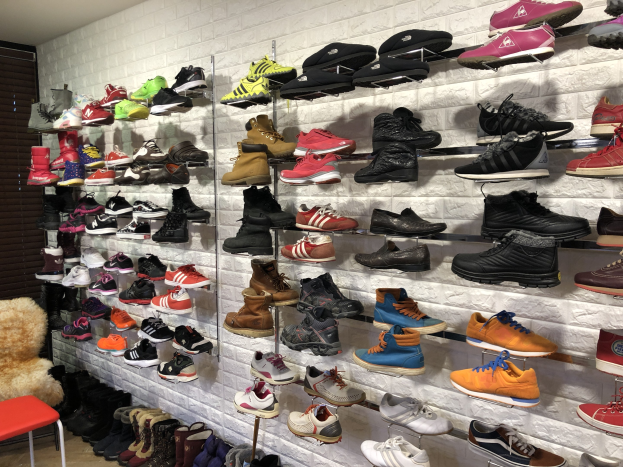 Ein Schuhgeschäft mit zahlreichen Schuhen auf den Ständern, einem Stuhl mit einem Teddybär auf der linken Seite und einer Wand im Hintergrund.