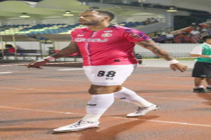 Ein Mann in einem pinken Hemd und weißen Shorts geht auf einem Fußballfeld, umgeben von sitzenden Zuschauern, einem Zaun, einem Dach mit Deckenleuchten und einer Wand.