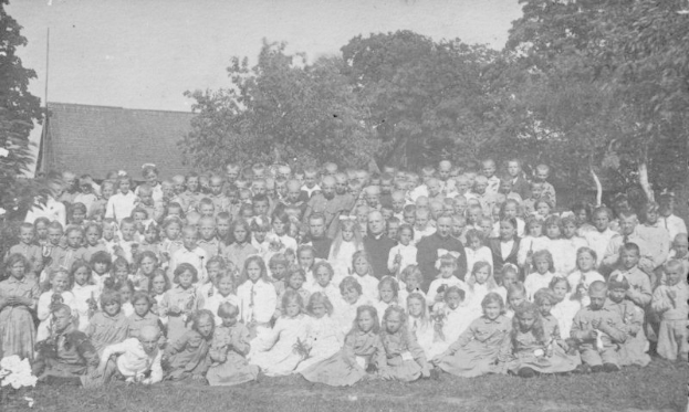 Schwarz-weißes Foto einer Gruppe von Kindern, die vor einem Haus mit Bäumen und einem klaren Himmel posieren.