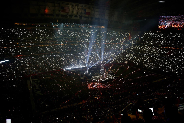 Eine gro├če Menge Menschen in einem Stadion bei Nacht, beleuchtet von hellen Lichtern, mit einem Bildschirm auf der rechten Seite, der wahrscheinlich die Ergebnisse der Super Bowl XLVII-Halbzeitenshow anzeigt.