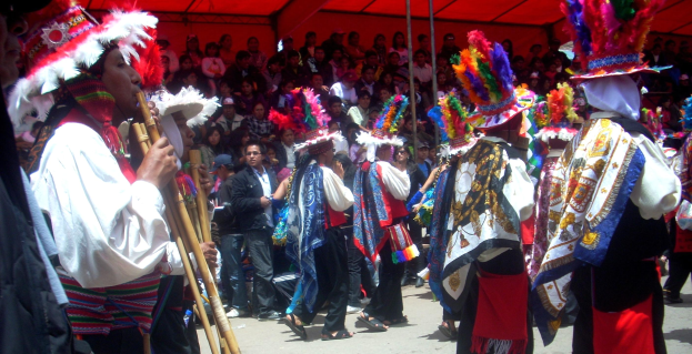 Eine Gruppe von Menschen in bunten Kostümen geht eine Straße entlang, mit einem Mann, der eine Flöte spielt, im Vordergrund und einer Menge, die unter einem Zelt sitzt, im Hintergrund auf einem peruanischen Festival.