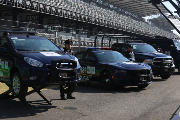 Eine Gruppe von Polizeiwagen vor einer Rennstrecke geparkt, mit uniformierten Beamten auf der Straße, mit einem Stadion und Maschendrahtzaun im Hintergrund.